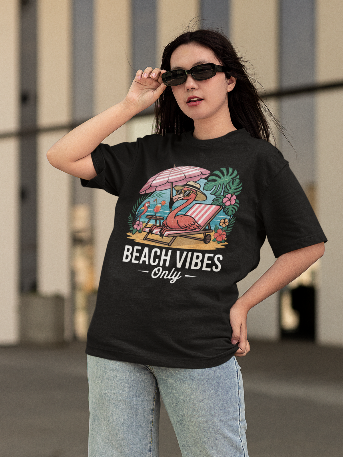 Mädchen mit Sonnenbrille. Sie trägt Jeanshose und ein schwarzes Shirt mit Aufdruck "Beach Vibes only" mit Flamingo unter Sonnenschirm.