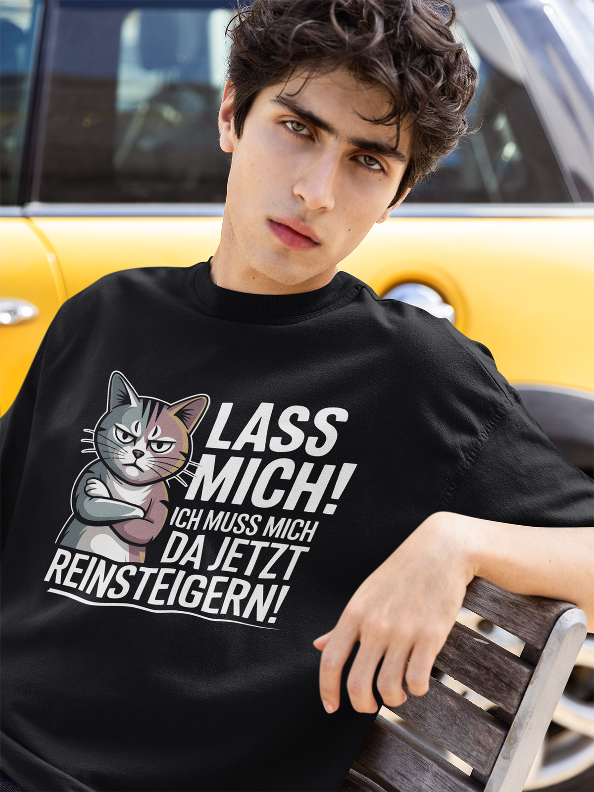 Junger Mann, sitzt auf einer Bank. Er trägt ein schwarzes T-Shirt mit Katzenmotiv und der Aufschrift "Lass mich! Ich muss mich da jetzt reinsteigern!". Im Hintergrund ein gelbes Auto.