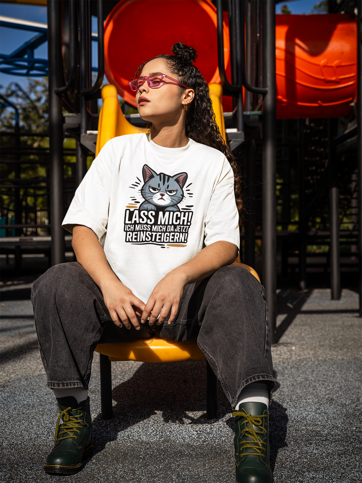 Junge Frau mit Brille sitzt auf gelber Rusche. Sie trägt eine graue Jeanshose und ein weißes Shirt mit großem Katzen-Motiv und den Spruch: "Lass mich! Ich muss mich da jetzt reinsteigern!"