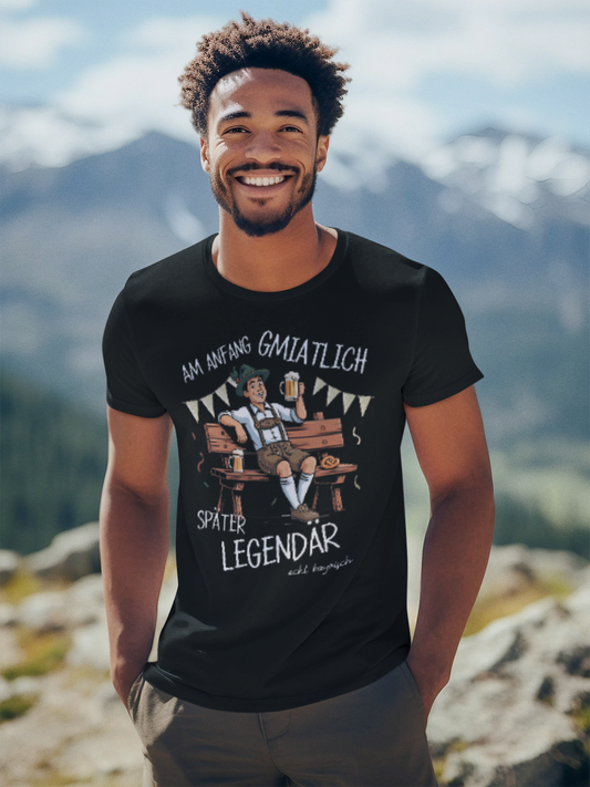 Mann trägt schwarzes T-Shirt mit Textmotiv „Am Anfang gmiatlich, später legendär“, Oberkörperansicht. Im Hintergrund Berge