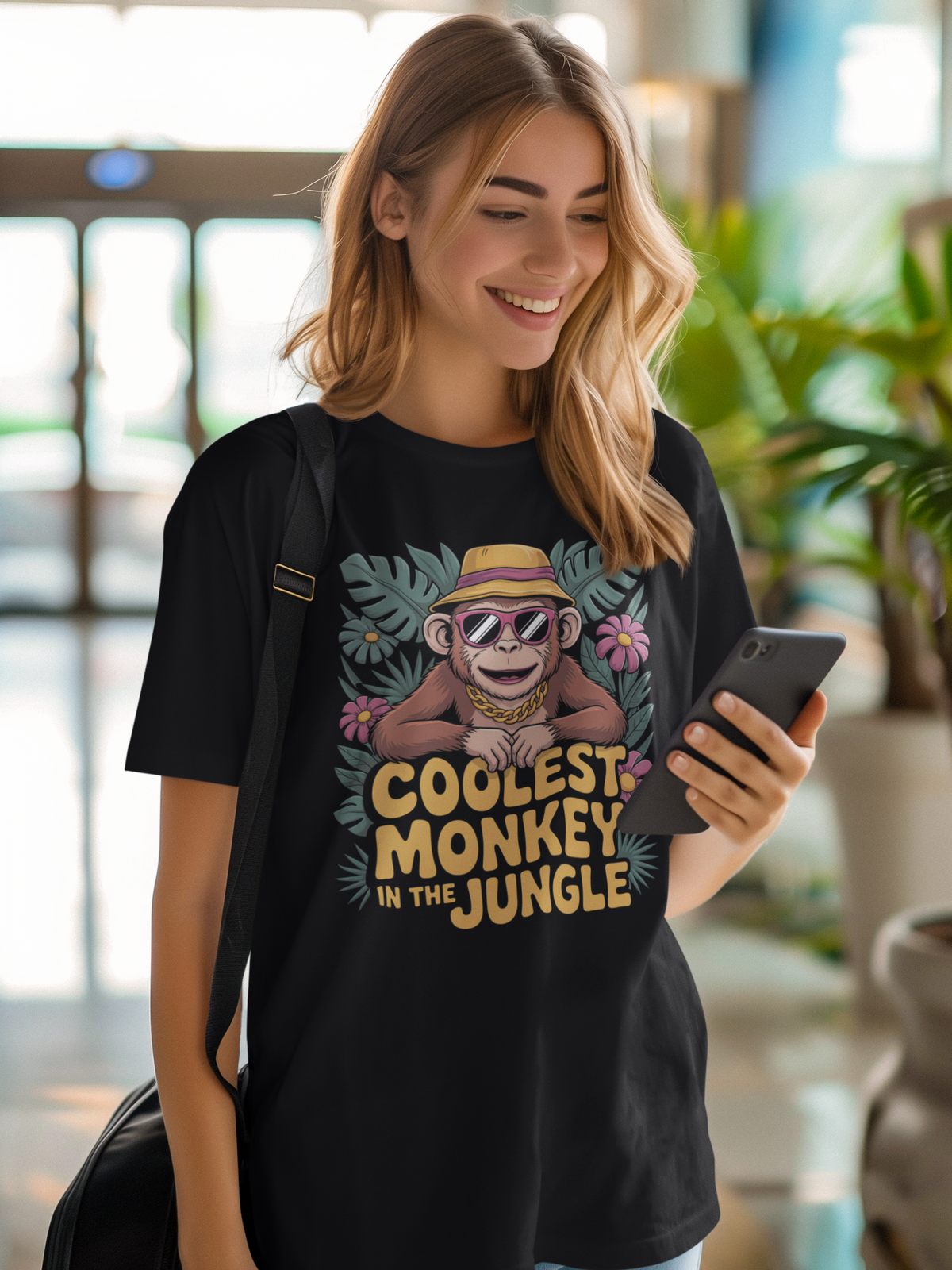 Junge blonde Frau, lachend mit Handy. Sie trägt ein schwarzes T-Shirt mit Affe mit Sonnenbrille und Hut, und Text „Coolest Monkey in the Jungle“