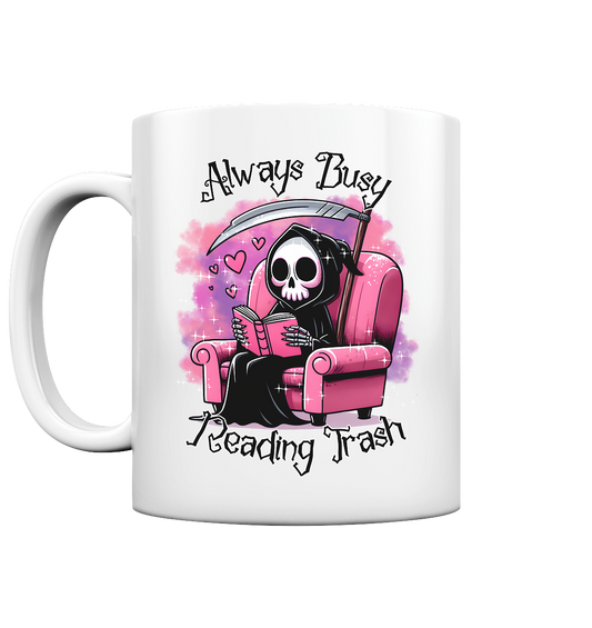 Weiße Tasse mit Motiv: Lesender süßer Sensemann auf rosa Sessel. Comic-Stil. Schriftzug: Always Busy Reading Trash.