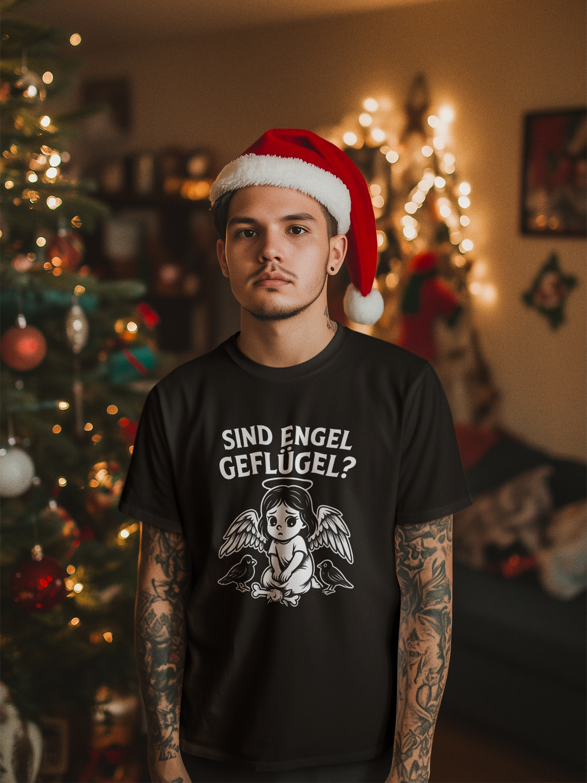 Junger Mann mit Weihnachtsmütze, im Hintergrund verschwommen ein Weihnachtsbaum. Er trägt ein schwarzes T-Shirt mit frechem Engel im Gothic-Comic-Stil und Schriftzug „Sind Engel Geflügel?“