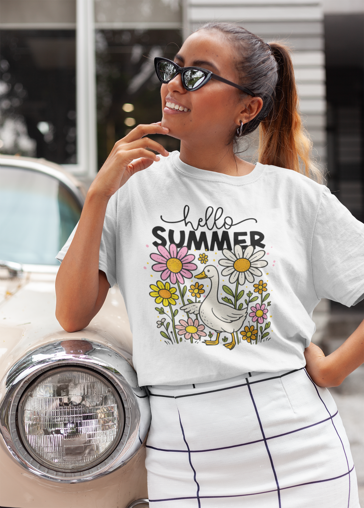 Frau lehnt lässig an einem Auto und trägt ein weißes T-Shirt mit Motiv: Gans im Blumenbeet und Schriftzug „Hello Summer“