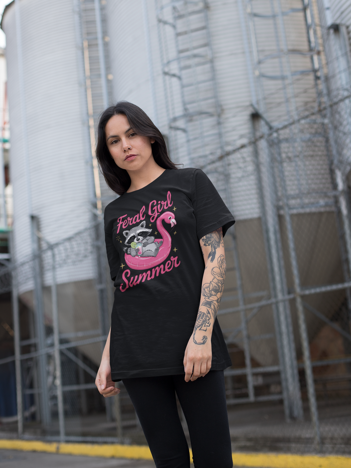 Mädchen trägt schwarzes T-Shirt mit Waschbär auf pinkem Flamingo-Schwimmreifen, Cocktail in der Pfote und Schriftzug „Feral Girl Summer“