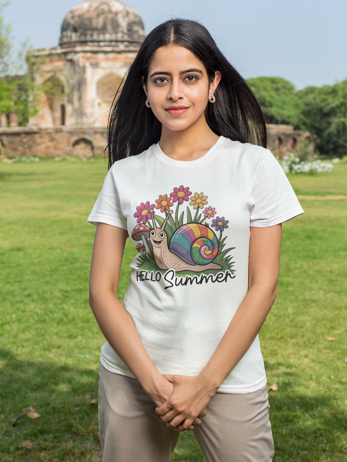 Junges Müdchen mit langen schwarzen Haaren trägt ein weißes T-Shirt mit süßem Comic-Motiv: kleine Schnecke im Blumenbeet, umgeben von Blüten, Text „Hello Summer“. Das Mädchen steht auf einer Wiese, mit Gebäude im Hintergrund.
