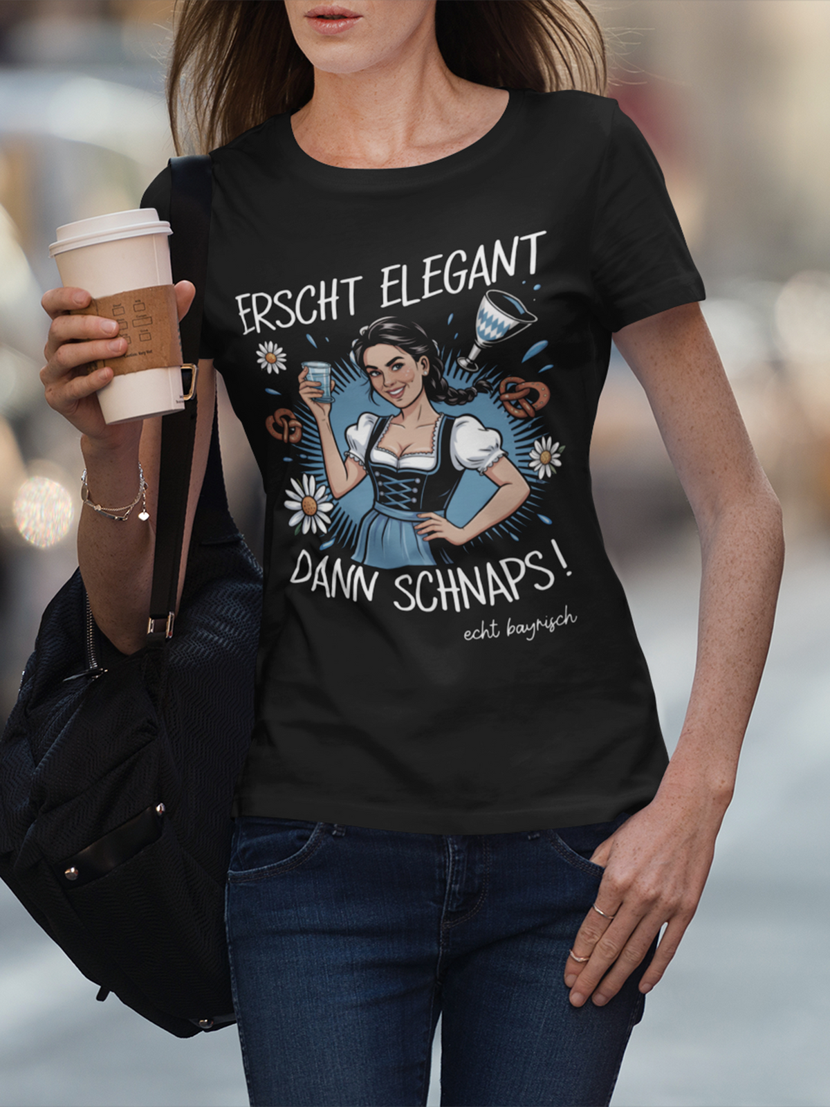 Frau trägt schwarzes T-Shirt mit Dirndl-Motiv und Spruch „Erscht elegant dann Schnaps“, Oberkörperansicht. Sie hält einen Kaffeebecher in der rechten Hand