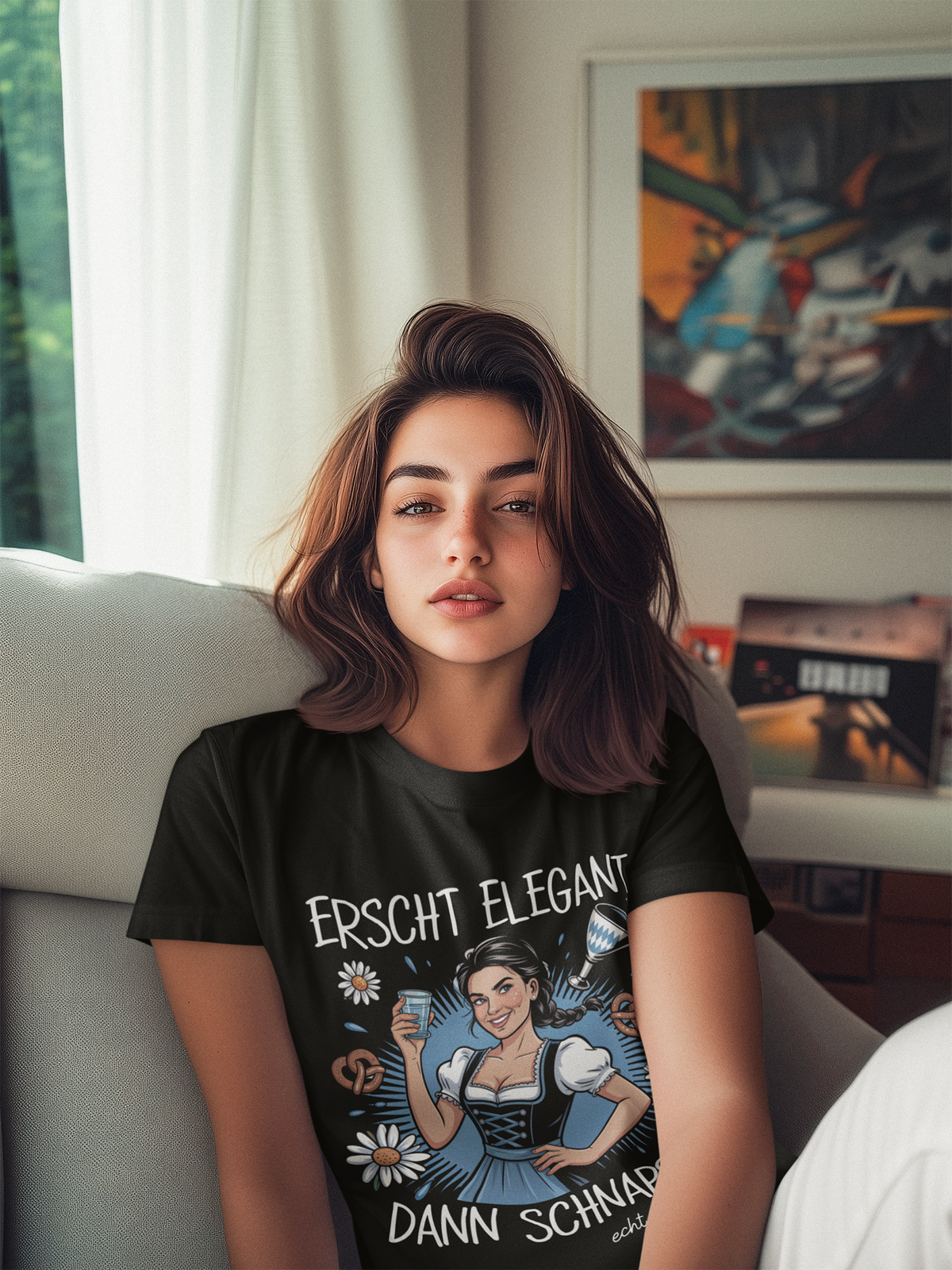 Frau sitzt auf Sofa, sie trägt schwarzes T-Shirt mit Dirndl-Motiv und Spruch „Erscht elegant dann Schnaps“, Oberkörperansicht