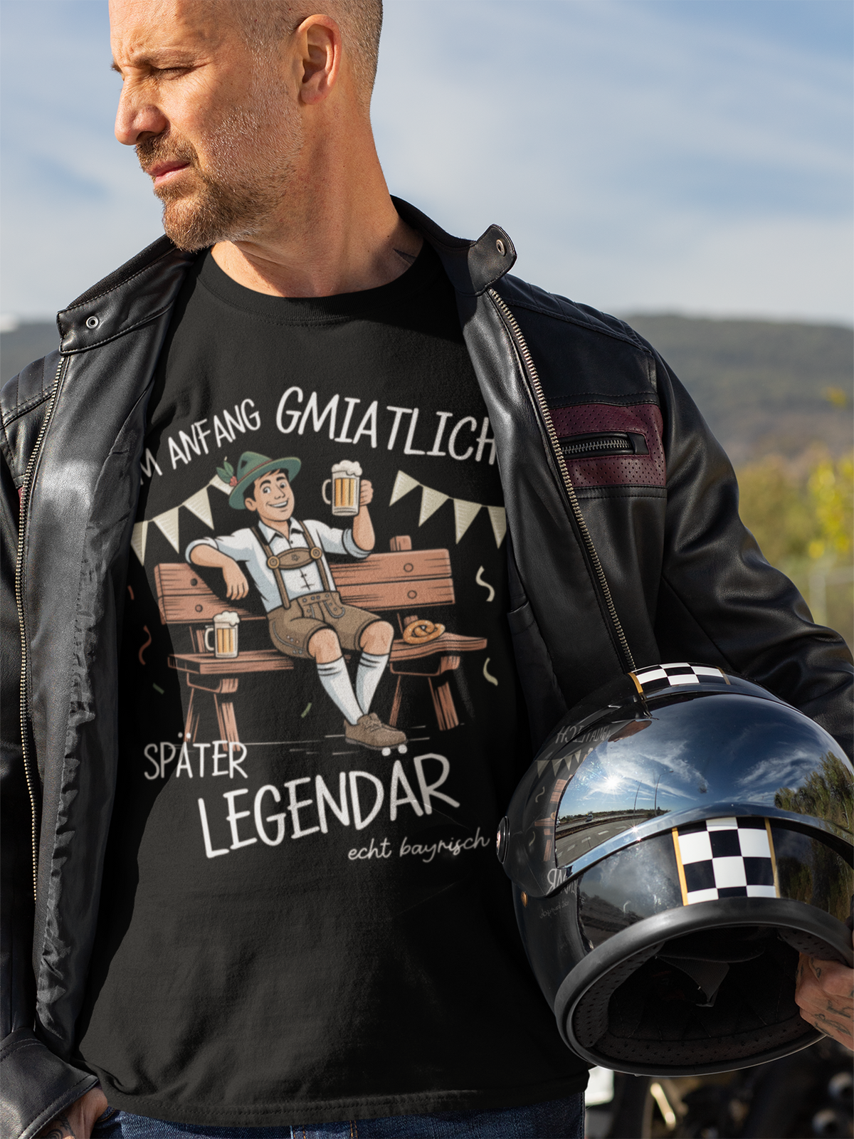 Mann mit Lederjacke und Helm unterm Arm. Er trägt schwarzes T-Shirt mit Textmotiv „Am Anfang gmiatlich, später legendär“, Oberkörperansicht