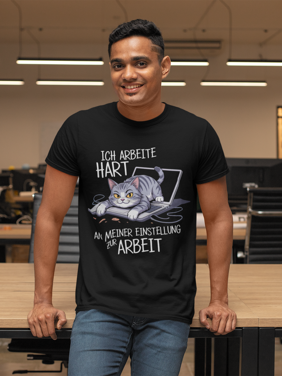 Mann, lehnt am Schreibtsich, er trägt schwarzes T-Shirt mit genervter Büro-Katze und frechem Spruch, Oberkörperansicht
