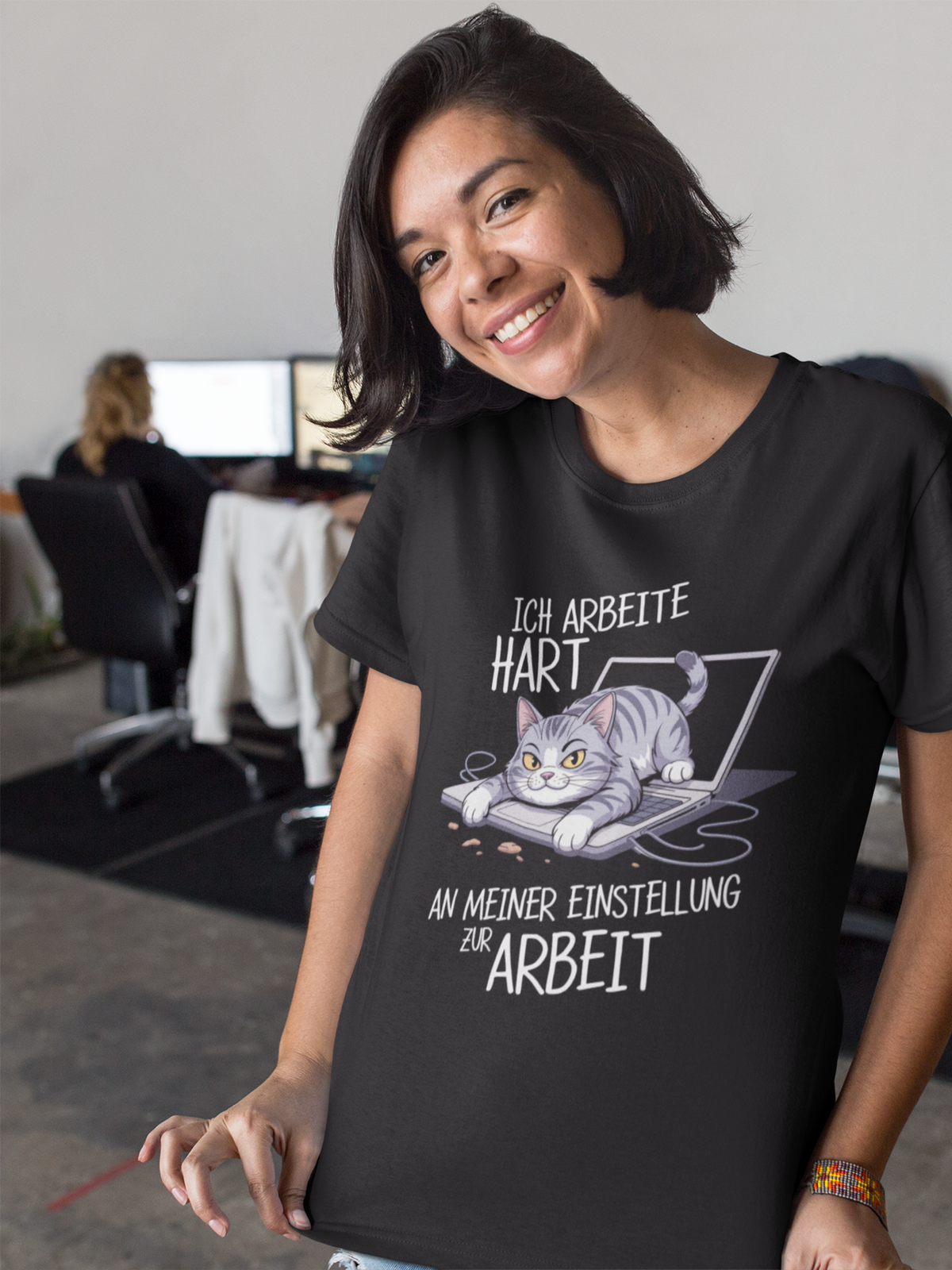 Junge Frau im Büro, zeigt ihr T-Shirt mit Katzen-Motiv und Spruch "Ich arbeite hart". Im Hintergrund Büro.