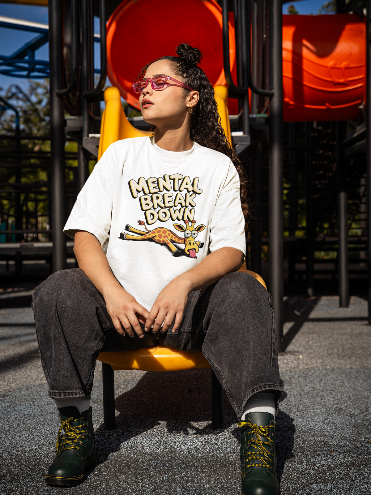 Junge Frau, sitzend auf Rutschbahn, trägt weißes T-Shirt mit Giraffe und dem Text „Mental Break Down“ (Ganzkörperaufnahme)