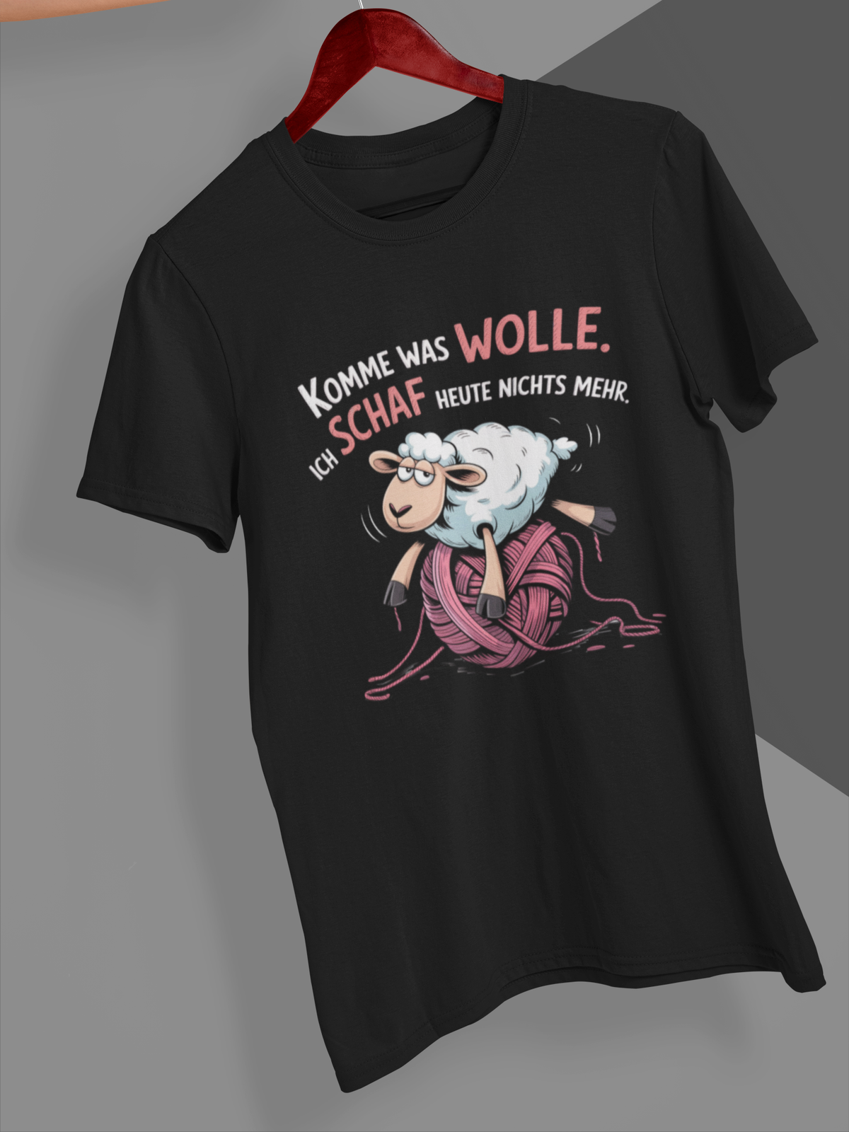 Schwarzes T-Shirt auf Kleiderbügel mit Schaf auf Wollknäuel und dem Spruch „Komme was Wolle. Ich Schaf heute nichts mehr“