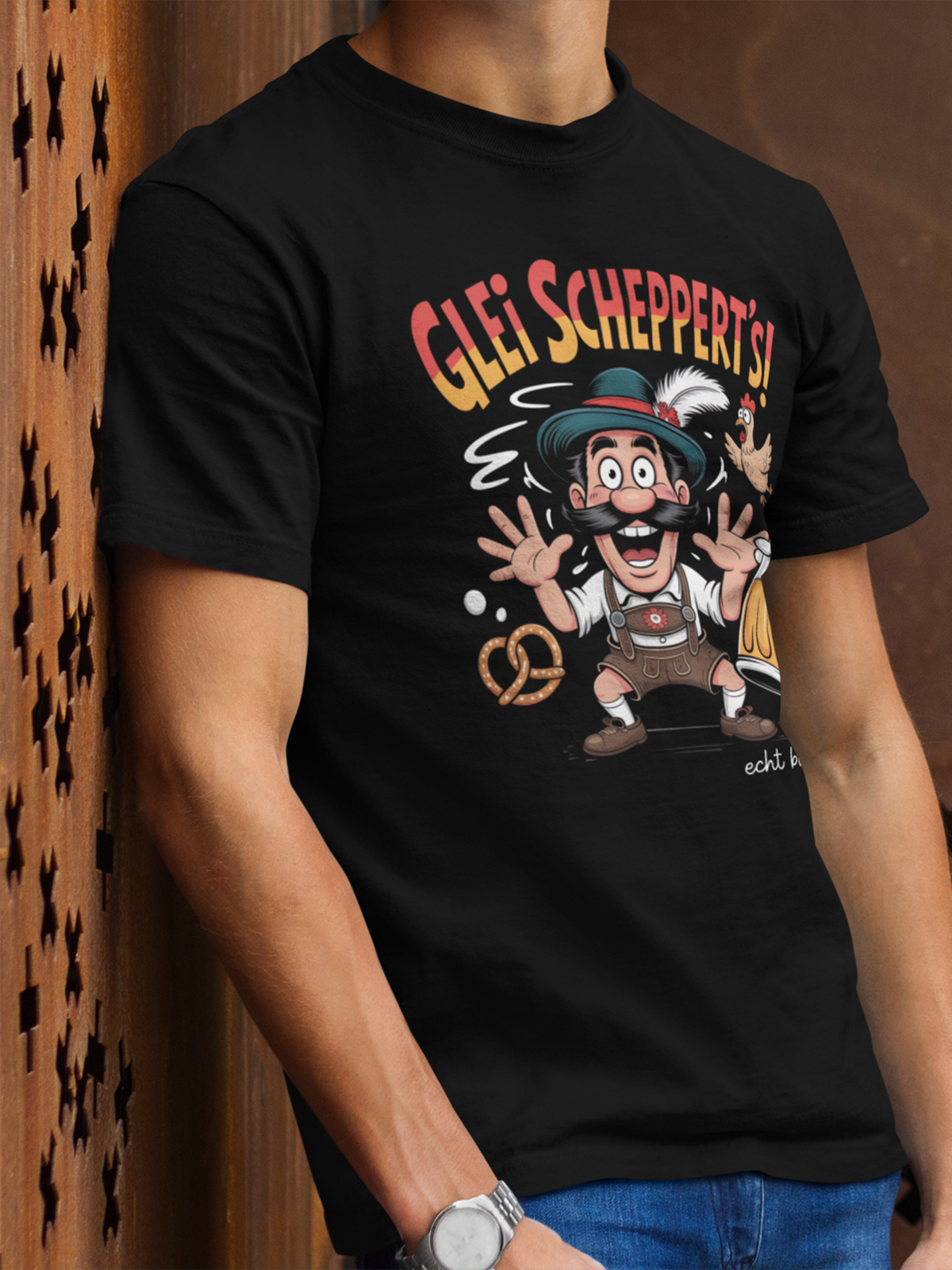 Mann Tattoos trägt ein schwarzes T-Shirt mit Aufschrift "Glei schppert´s. Echt bayrisch" und Bayern-Motiv: Mann in Lederhose und Masskrug. Oberkörperansicht