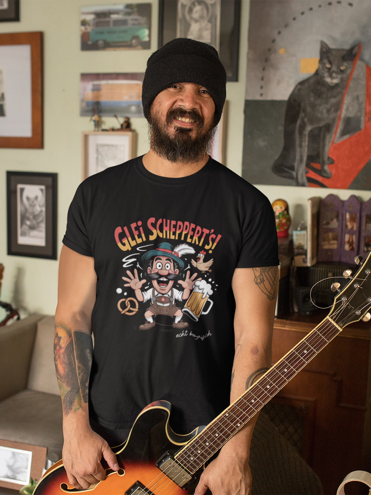 Mann mit Bart und Tattoos trägt ein schwarzes T-Shirt mit Aufschrift "Glei schppert´s. Echt bayrisch" und Bayern-Motiv: Mann in Lederhose und Masskrug. Unten im Bild eine E-Gitarre, im Hintergrund Bilder an der Wand.