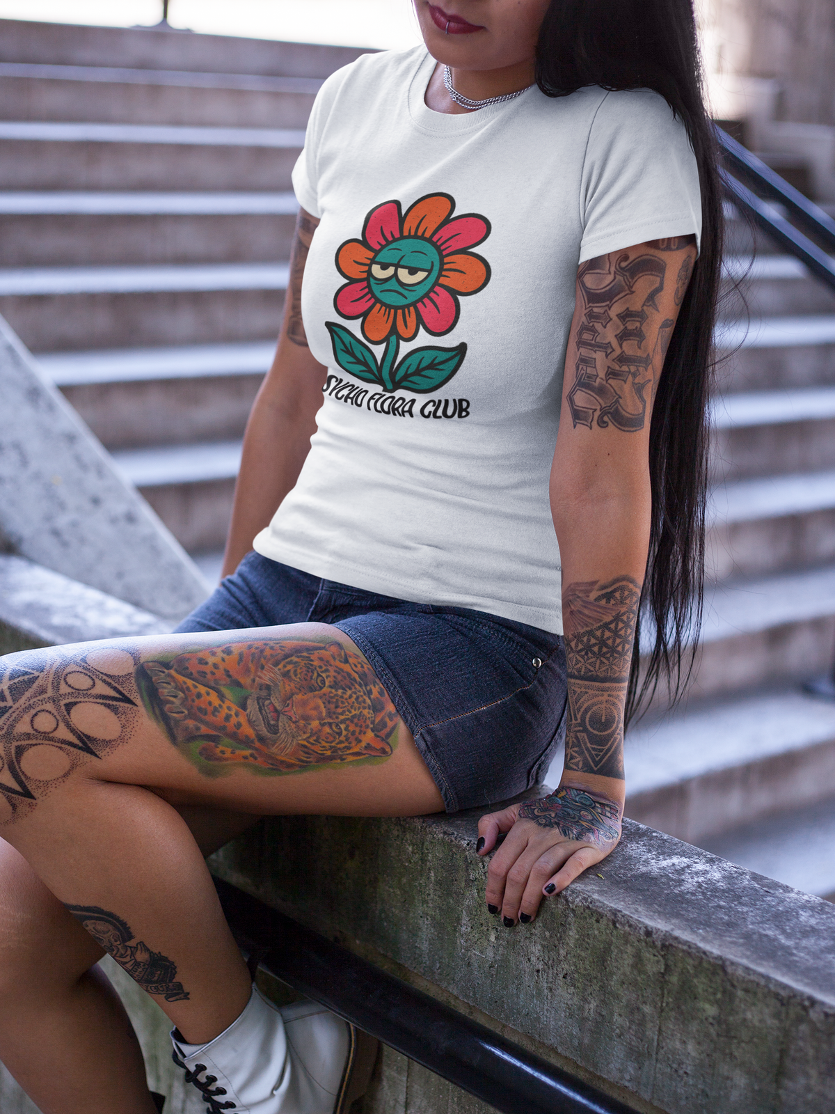 Junge tättowierte Frau trägt eine kurze Hose und ein Weißes T-Shirt mit Retro-Blume mir Augen und Schriftzug „Psycho Flora Club“. Sie sitzt auf einer Mauer.