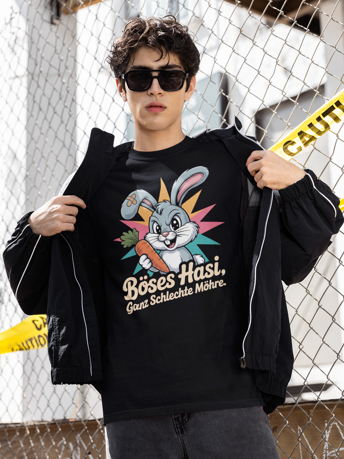 Junger Mann mit Sonnebrille, trägt schwarze offene Jacke und schwarzes Shirt mit Hase und Möhre (Oberkörperaufnahme), vor Zaun