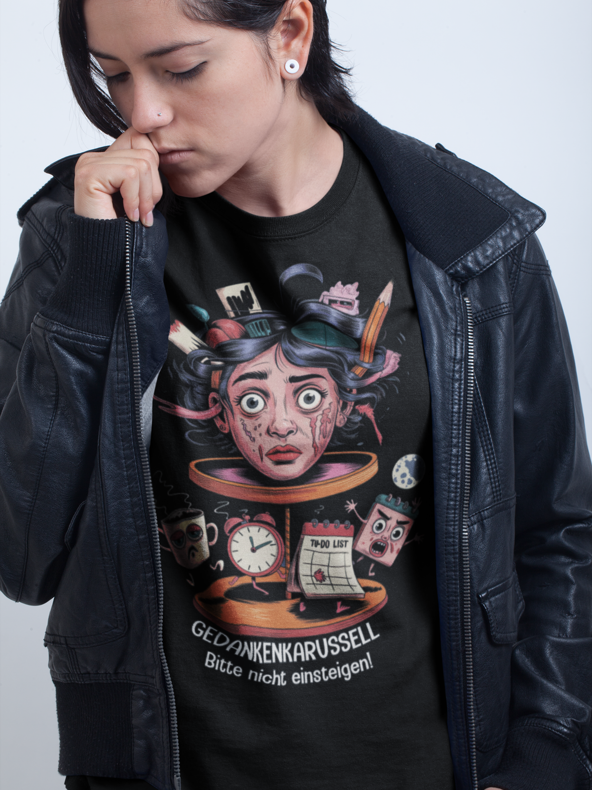 Junge Frau mit offener schwarzer Lederjacke. Darunter trägt sie ein schwarzes T-Shirt mit surrealistischem Kopf-Karussell-Motiv und weißem Text „Gedankenkarussell – Bitte nicht einsteigen“