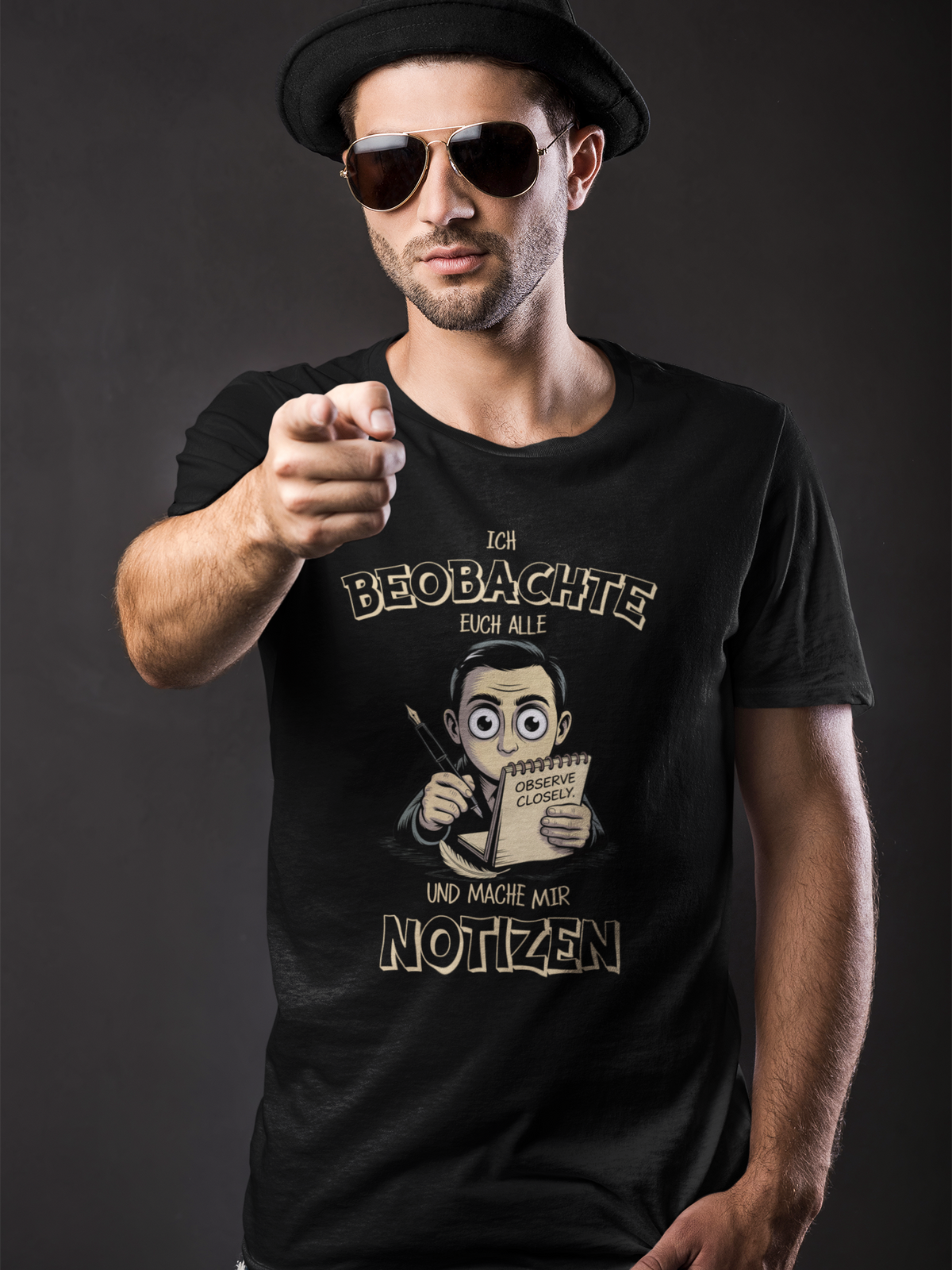 Mann mit Sonnenbrille, zeigt auf die Kamera, er trägt schwarzes Shirt mit dem Spruch „Ich beobachte euch alle und mache mir Notizen“ (Oberkörperaufnahme)
