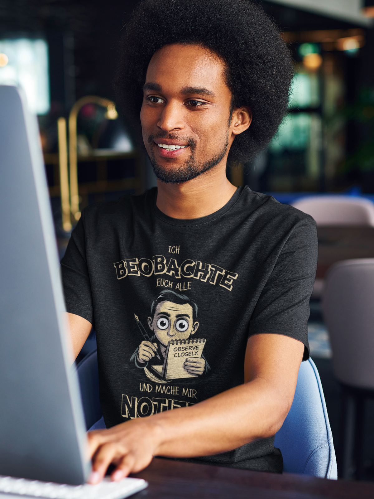 Junger schwarzer Mann, sitzend, am Bildschirm, er trägt schwarzes Shirt mit dem Spruch „Ich beobachte euch alle und mache mir Notizen“ (Oberkörperaufnahme)