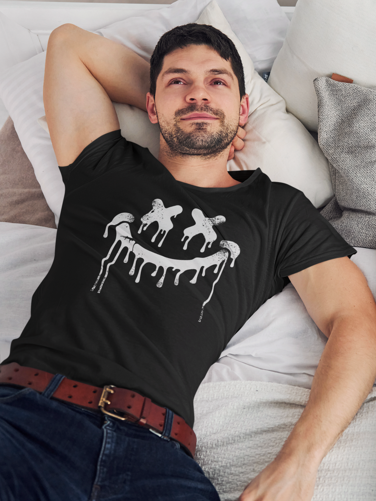 Junger Mann, liegend auf Bett, trägt ein schwarzes T-Shirt mit Smilie-Gesicht im Graffiti-Look. Oberkörperansicht.