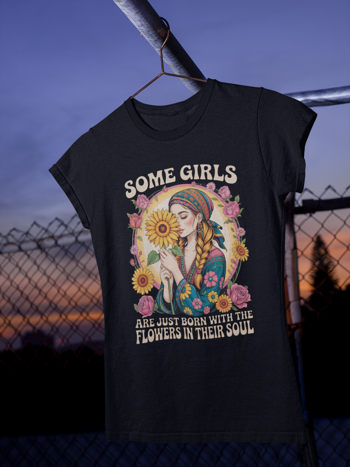 Schwarzes Ladies T-Shirt mit dem Text „Some Girls are just born with the flowers in their soul“ und floralem Design, hängt auf Kleiderbügel. Im Hintergrund ein Zaun.