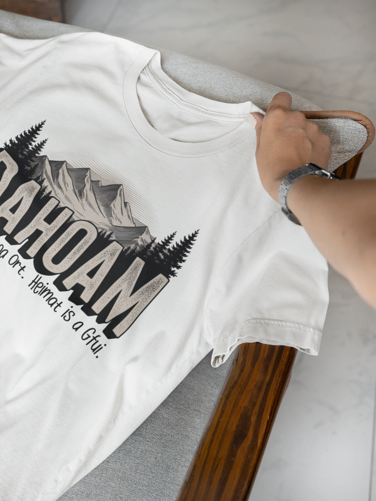 Weißes T-Shirt, liegend, Teilansicht mit Hand,  mit dem Spruch „Dahoam. Heimat is koa Ort. Heimat is a Gfui“ und Bergmotiv
