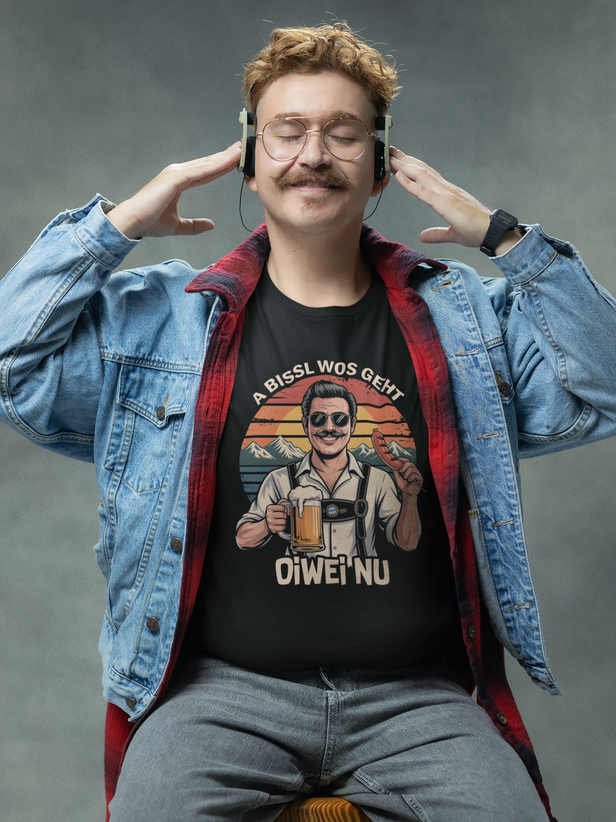 Mann, sitzend, mit offener Jeansjacke, trägt schwarzes T-Shirt mit Retro-Motiv „A bissl wos geht oiwei nu“ (Oberkörper)