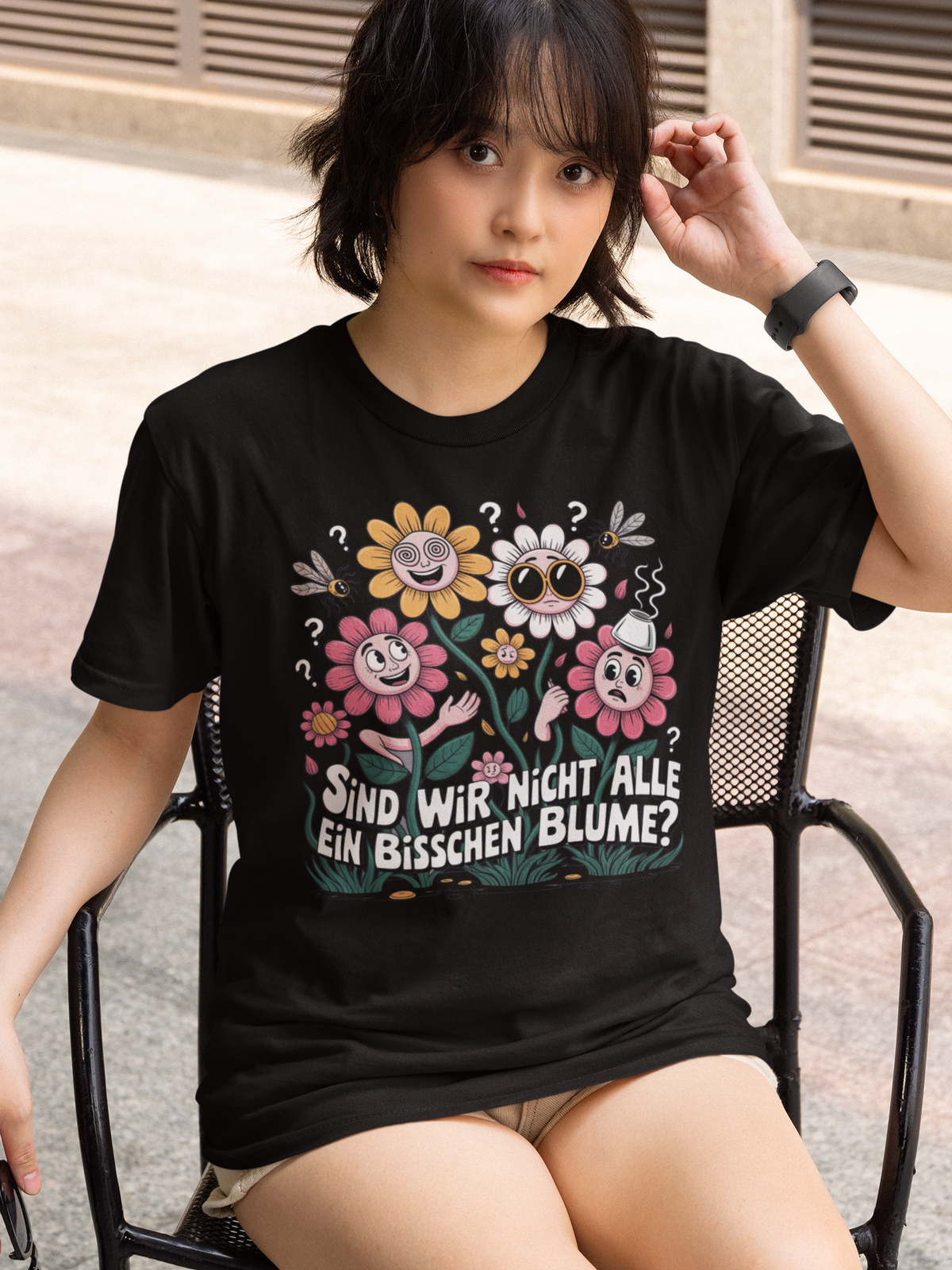 Junge Frau, auf Stuhl sitzend, trägt schwarzes T-Shirt mit buntem Blumenmotiv und Spruch „Sind wir nicht alle ein bisschen Blume?“ (Oberkörper)