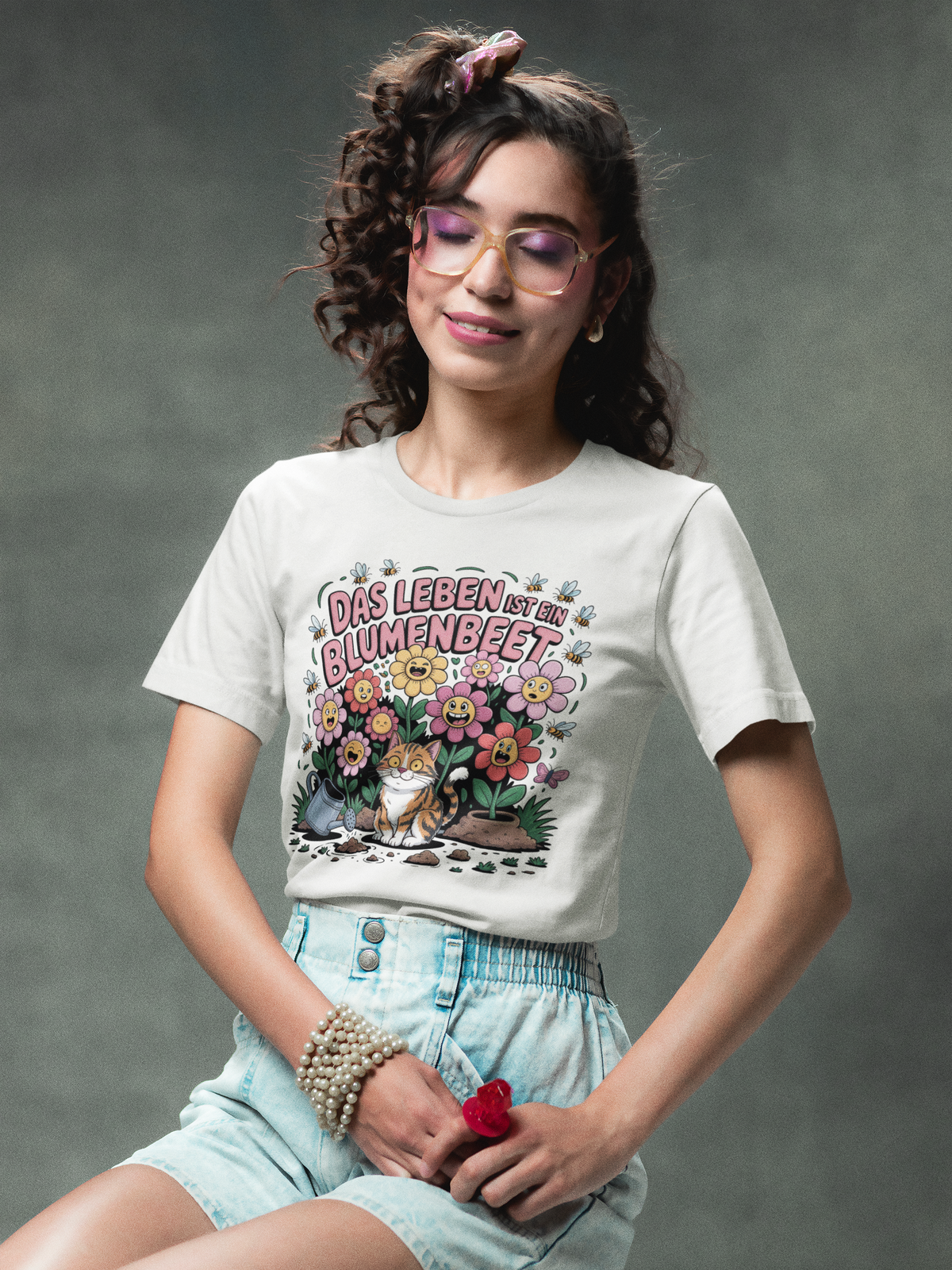 JUnge Frau, sitzend, mit Brille, trägt weißes T-Shirt mit Motiv „Das Leben ist ein Blumenbeet“ und Katze (Oberkörper)

