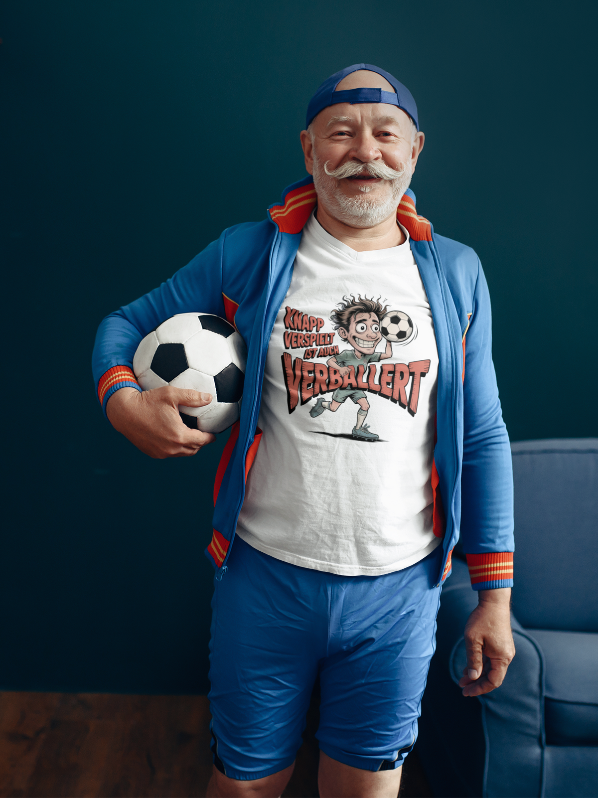 Älterer Mann, Fußball unterm Arm, blaue offene Trainingsjacke, darunter trägt er ein weißes T-Shirt mit Fußballspruch „Knapp verspielt ist auch verballert“ (Oberkörper)

