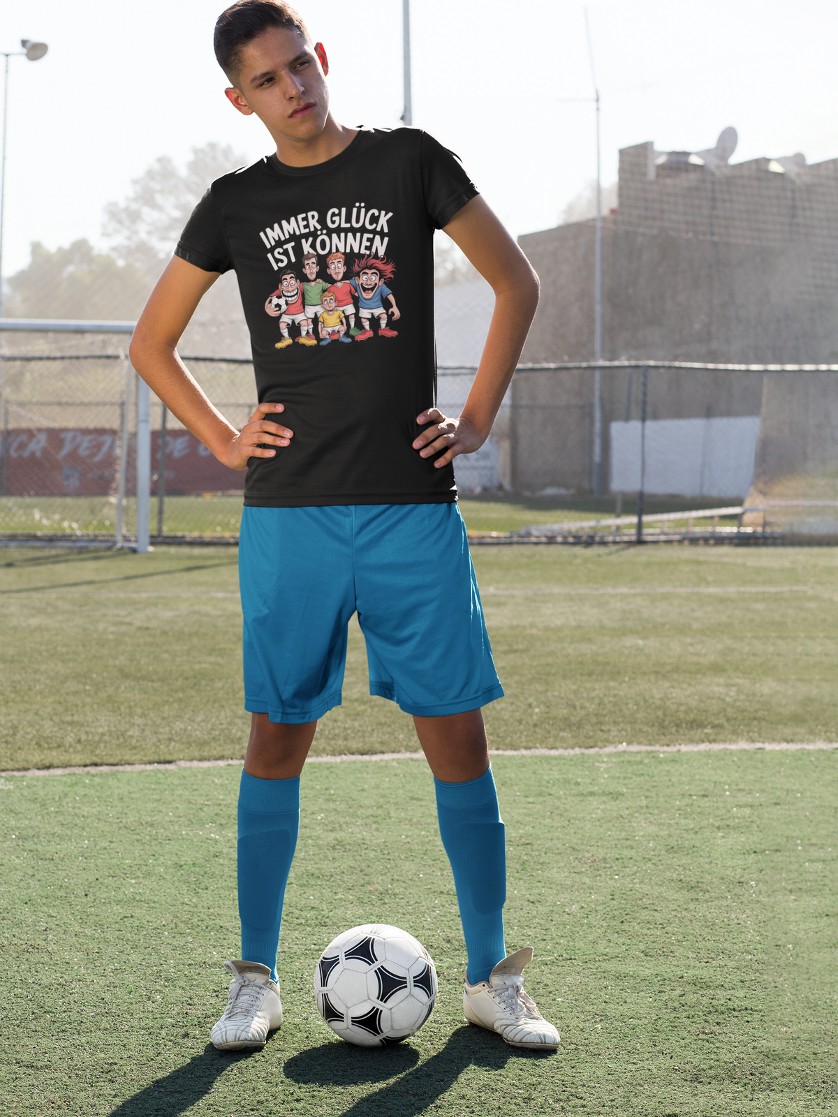 Fußballspieler auf rasen mit Ball, blaue Hose und schwarzes Shirt  mit Spruch „Immer Glück ist Können“ und Fußballteam-Motiv (Ganzkörper)