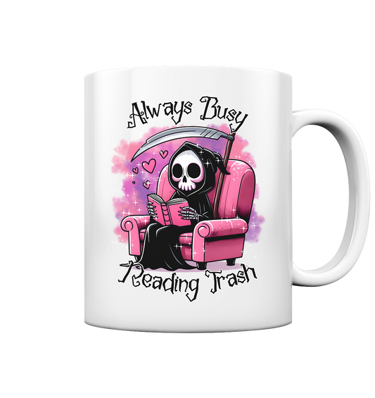 Weiße Tasse mit Motiv: Lesender süßer Sensemann auf rosa Sessel. Comic-Stil. Schriftzug: Always Busy Reading Trash.