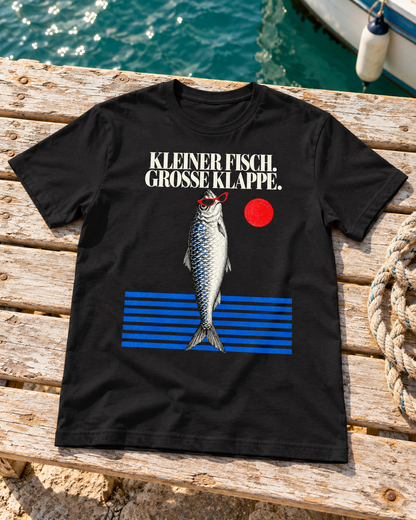 KI-Szenenbild: Schwarzes T-Shirt „Kleiner Fisch, große Klappe“ liegt neben einem Tau auf einer Holzbank am Hafen