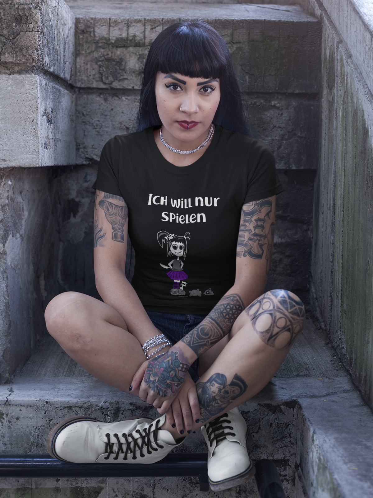 Mödchen, sitzend mit verschränkten Beinen trägt schwarzes T-Shirt mit weißem Schriftzug „Ich will nur spielen“ und frechem Gothic Girl auf der Vorderseite